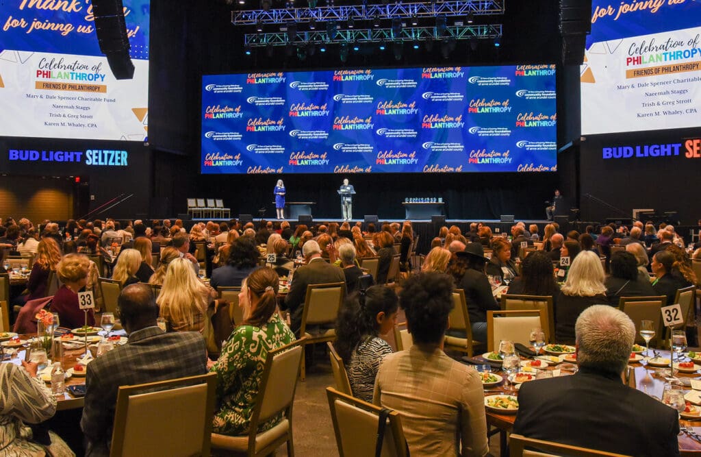 A packed ballroom at The Hall at Live! Casino & Hotel Maryland during the Community Foundation of Anne Arundel County's Celebration of Philanthropy Awards Luncheon. Hundreds of guests are seated at numbered round tables for a formal luncheon while two presenters stand at the podium on stage.