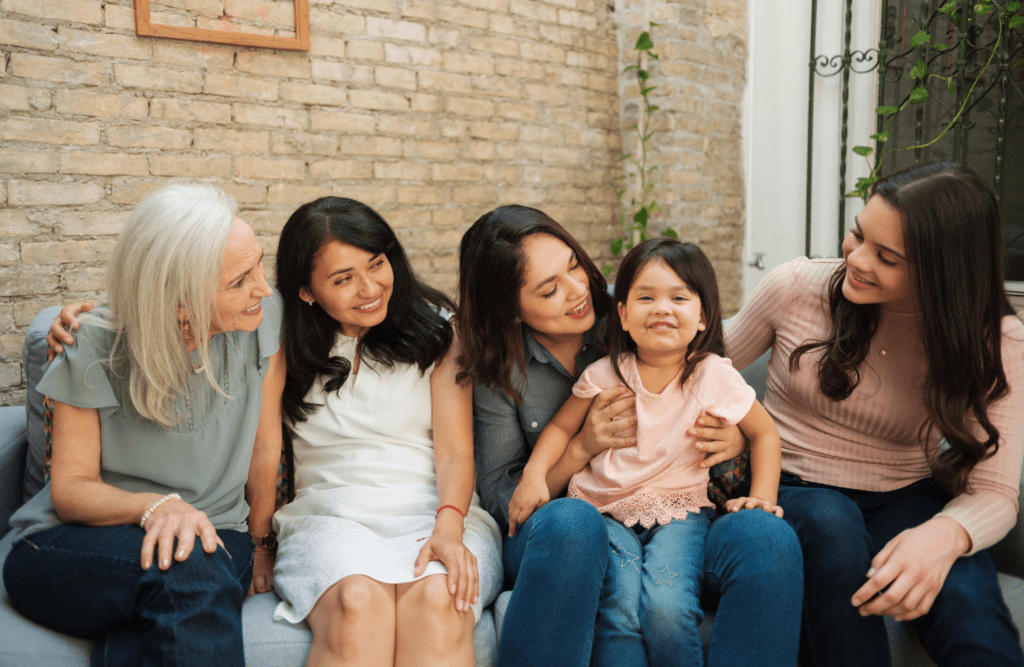 Women of different generations sit together on a sofa laughing and smiling, with a young girl in a pink shirt seated in the middle, all gathered in a warm…Four women of different generations sit together on a sofa laughing and smiling, with a young girl in a pink shirt seated in the middle, all gathered in a warm, brick-walled outdoor setting.