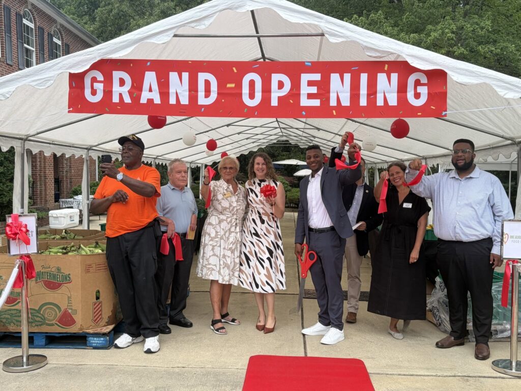 Community members and officials cut a ribbon under a tent with a red 'Grand Opening' banner, celebrating the launch of a community market by Anne Arundel County Food Bank and The Miracle Temple Inc. Boxes of fresh watermelons and corn are displayed in the foreground.