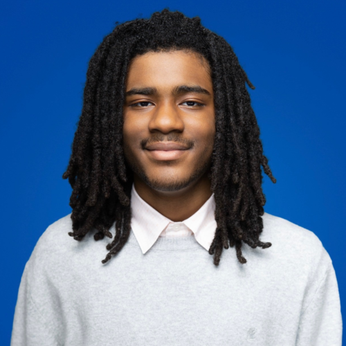 Andre Manning Jr., Young Volunteer of the Year 2026: Headshot of Andre Manning Jr. against a solid blue background, wearing a light gray sweater over a white collared shirt.