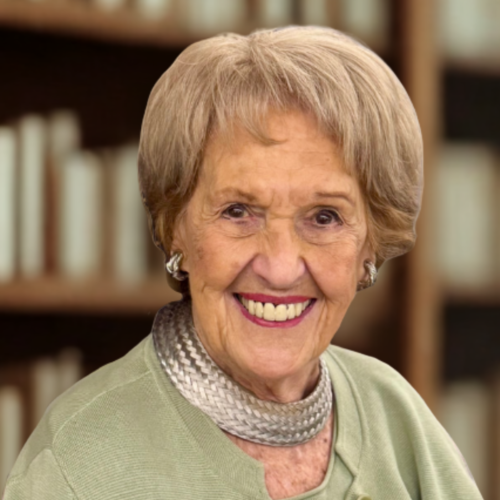Portrait of Anna E. Greenberg, an older woman with short blonde hair, smiling and wearing a green cardigan and a silver woven choker necklace, with a blurred bookshelf in the background.