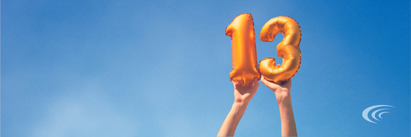 Two hands raised against a blue sky, holding orange balloon numbers spelling "13."