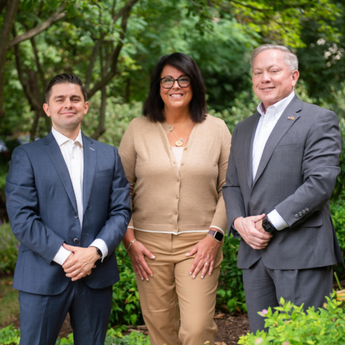 Billy Hufnell, Amy Francis, and Bill Hufnell of Modera standing together outdoors in front of green foliage, dressed in business attire.